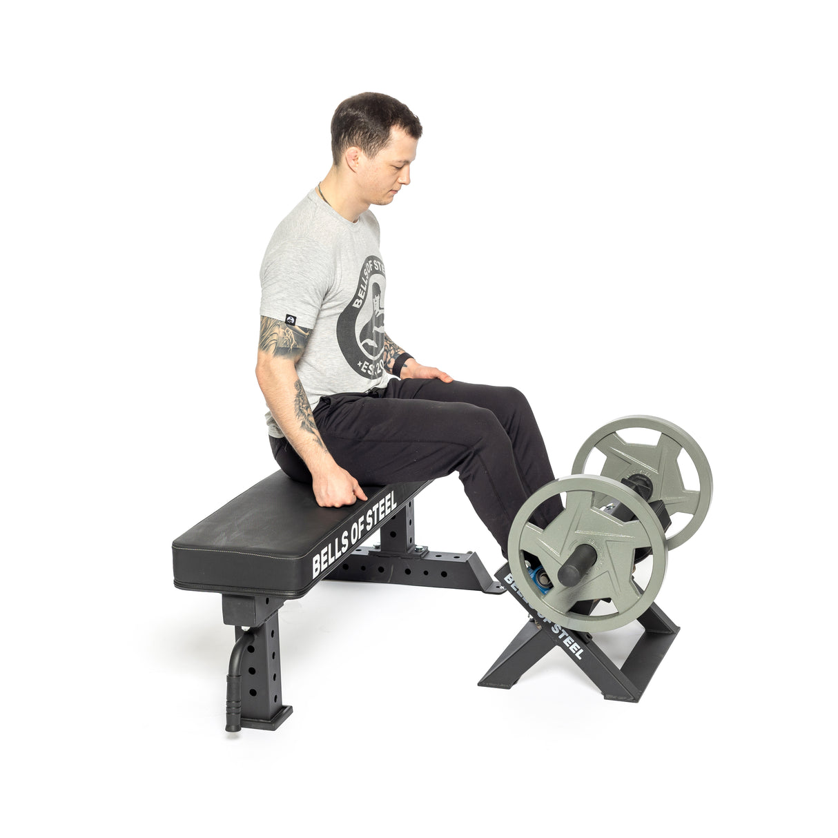 A person uses the Bells of Steel Tibialis Trainer Machine, performing wrist curls with a barbell on a stand, wearing a gray t-shirt and black pants. They focus intently on their strength training routine to achieve balanced muscle development.