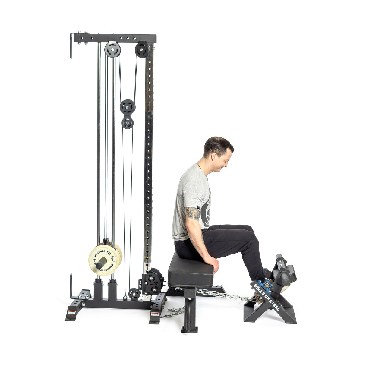 A person uses a Bells of Steel Tibialis Trainer Machine for strength training, facing right with feet on the foot platform, wearing a gray shirt and black pants. The black equipment utilizes weights and pulleys to target the tibialis anterior muscles.