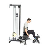 A person uses a Bells of Steel Tibialis Trainer Machine for strength training, facing right with feet on the foot platform, wearing a gray shirt and black pants. The black equipment utilizes weights and pulleys to target the tibialis anterior muscles.