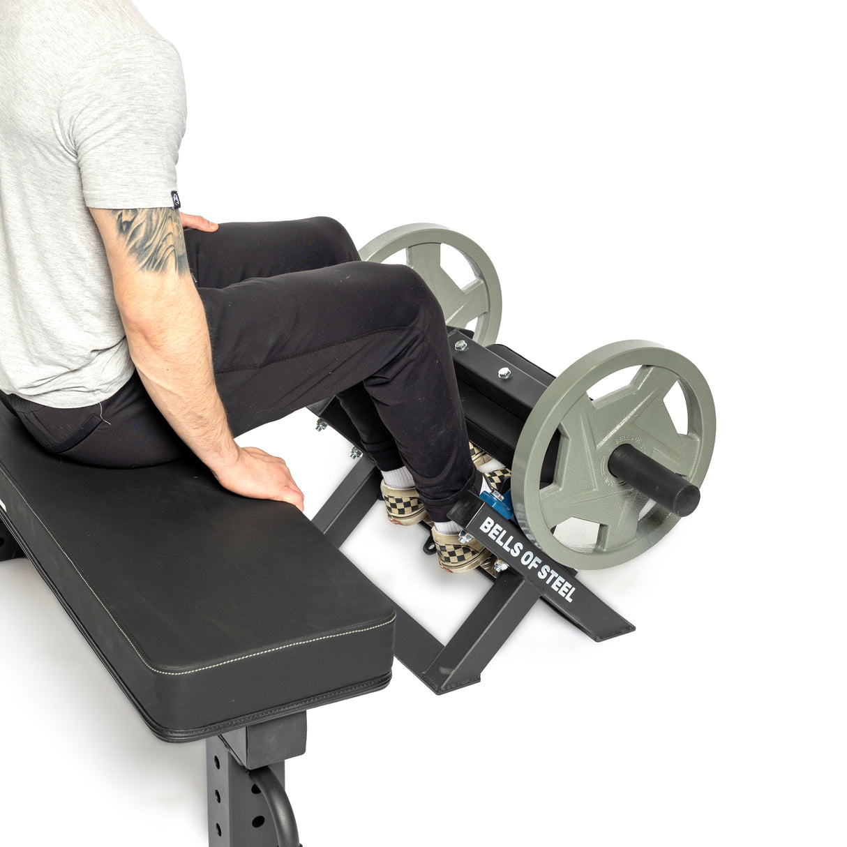 A person in a gray shirt and black pants sits on a workout bench, legs bent, using the "Bells of Steel" Tibialis Trainer Machine. The focus is on green weight plates, their tattooed arm, and the machine for effective strength training against a plain white backdrop.