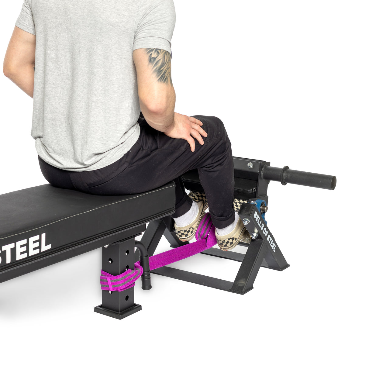 A person seated on a bench uses the Tibialis Trainer Machine by Bells of Steel for strength training. Wearing a gray shirt, black pants, and checkered shoes, their ankles are secured by a purple strap attached to the frame, fully engaging their tibialis anterior muscles.