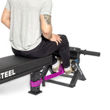 A person seated on a bench uses the Tibialis Trainer Machine by Bells of Steel for strength training. Wearing a gray shirt, black pants, and checkered shoes, their ankles are secured by a purple strap attached to the frame, fully engaging their tibialis anterior muscles.