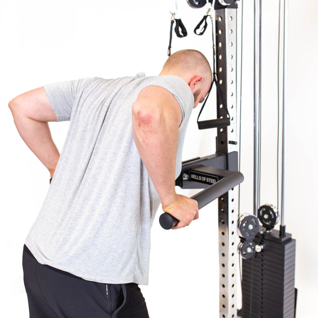 A person in a gray shirt and black pants is focused on performing tricep dips with precision using adjustable handles on the Bells of Steel Cable Tower.