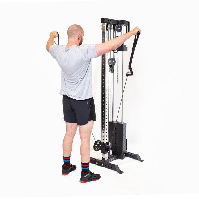 Someone in a gray t-shirt and black shorts is exercising with a Bells of Steel Cable Tower, pulling the adjustable handles upwards with both hands on a white background.