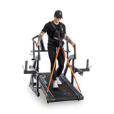 A person on a sled treadmill, in a black shirt and cap, wears a Bells of Steel Weighted Vest. They grip the side rails while standing on the belt. The KEISER equipment features a strong metal frame with harnesses connected to orange ropes for an intense workout.