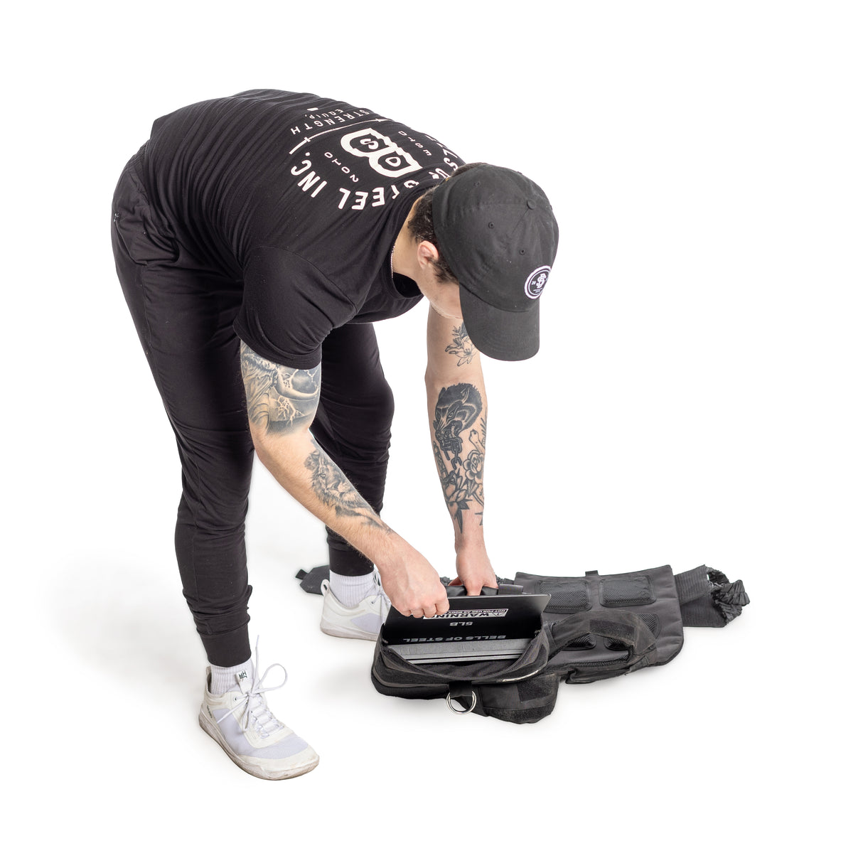 A tattooed individual in black attire and cap places their laptop into a black backpack, along with Bells of Steel USA Weighted Vest and Plates, against a stark white background, hinting at future cardio gains.