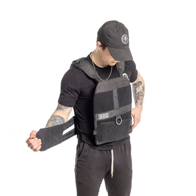 A person in a black outfit and cap focuses intently while adjusting a strap on a Bells of Steel Weighted Vest. Tattoos adorn their arms, set against a plain white background.