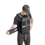 A person in a black cap and shirt adjusts their Bells of Steel USA Weighted Vest and Plates, showcasing tattooed arms against a stark white backdrop.