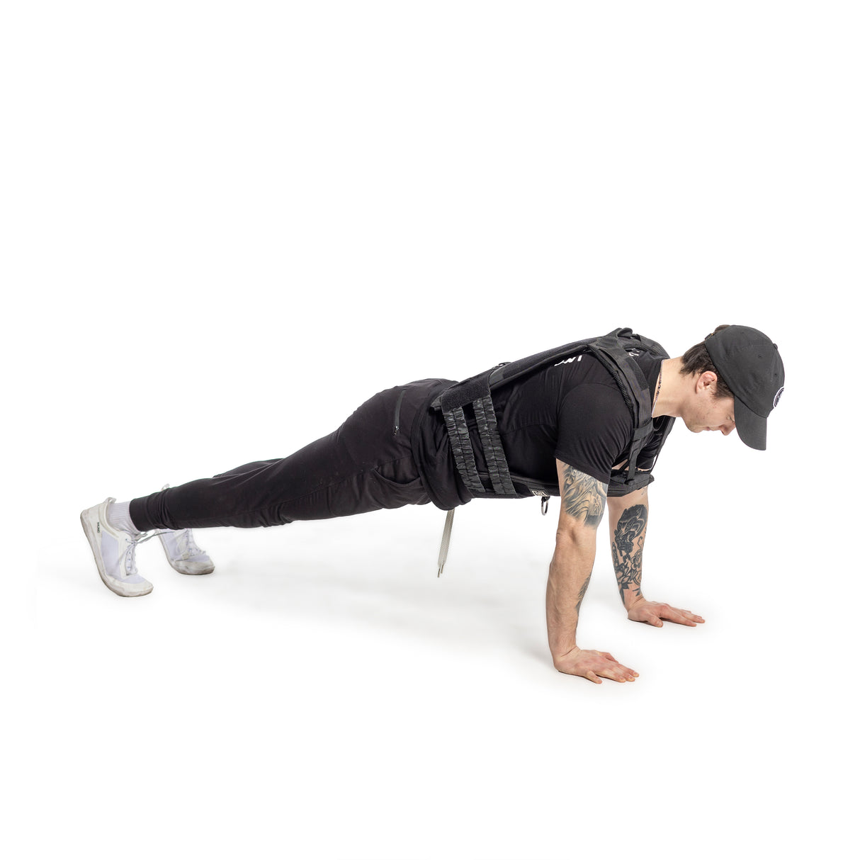 A person in a cap, black attire, and a Bells of Steel Weighted Vest does a plank on their hands and toes against a white backdrop.