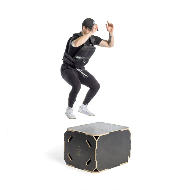 In a studio with a white backdrop, someone in workout attire and a cap performs a box jump while wearing a Bells of Steel Weighted Vest onto a sturdy black box labeled "20 exx.