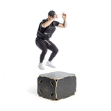 A person in a black Bells of Steel USA Weighted Vest and Plates, wearing a cap and white sneakers, is mid-air while jumping onto a black plyometric box against a plain white background.