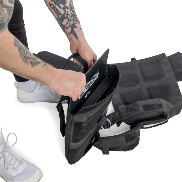 A person with tattoos slips a black rectangular object into the pocket of their Bells of Steel Weighted Vest. The adjustable vest complements their black pants and white sneakers against the plain white background.