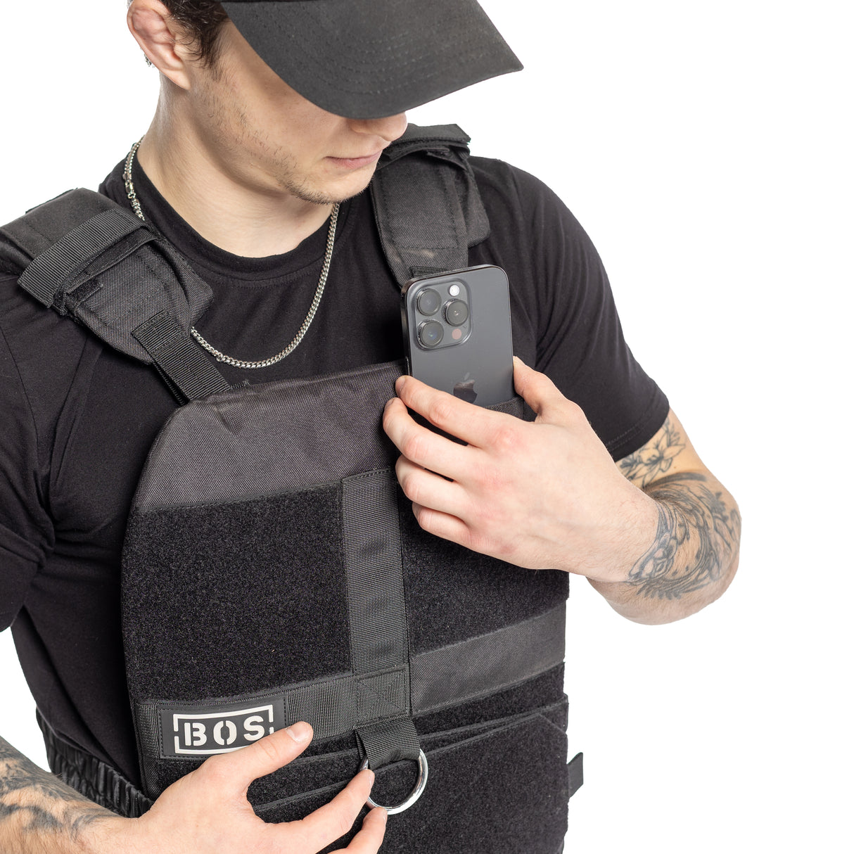 A person in a black tactical Bells of Steel Weighted Vest with training plates inserts an iPhone into the vest's pocket. Dressed in a black shirt and cap, their arms display tattoos. The adjustable vest features a D-ring and the "BOS" logo.