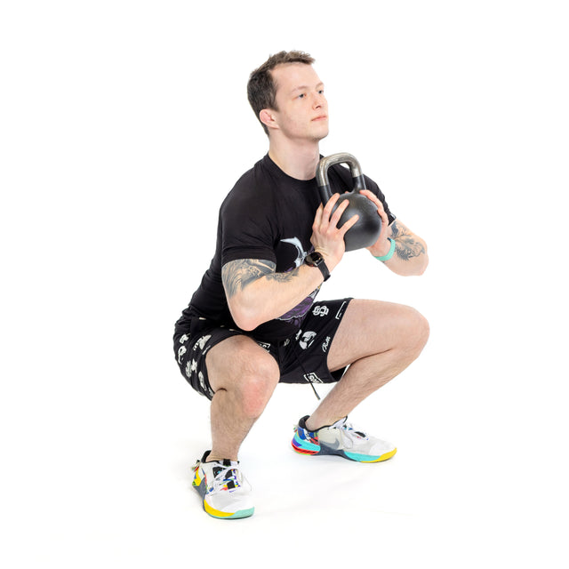 A person squats with a Mark Wildman Adjustable Kettlebell by Bells of Steel, capturing the Wildman Athletica spirit in a black shirt, patterned shorts, and colorful sneakers against a white backdrop.