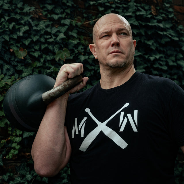 A bald man in a black T-shirt holds the Bells of Steel USA 48KG Extension Kit for Adjustable Kettlebell (Wildman Poison Pill) on his shoulder, standing before a green ivy-covered wall with a serious expression.