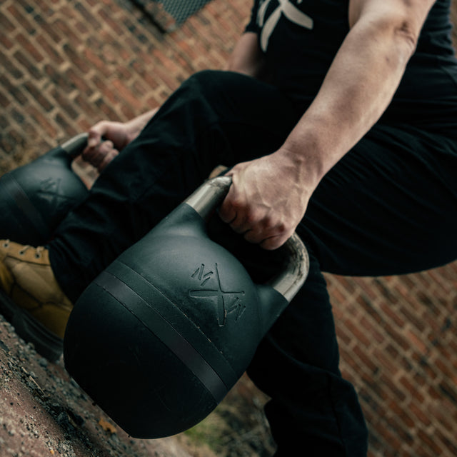 Wearing athletic gear, a person squats outdoors on rough ground, gripping the 48KG Extension Kit for Adjustable Kettlebell – Wildman Poison Pill by Bells of Steel USA; a brick wall is seen in the background.