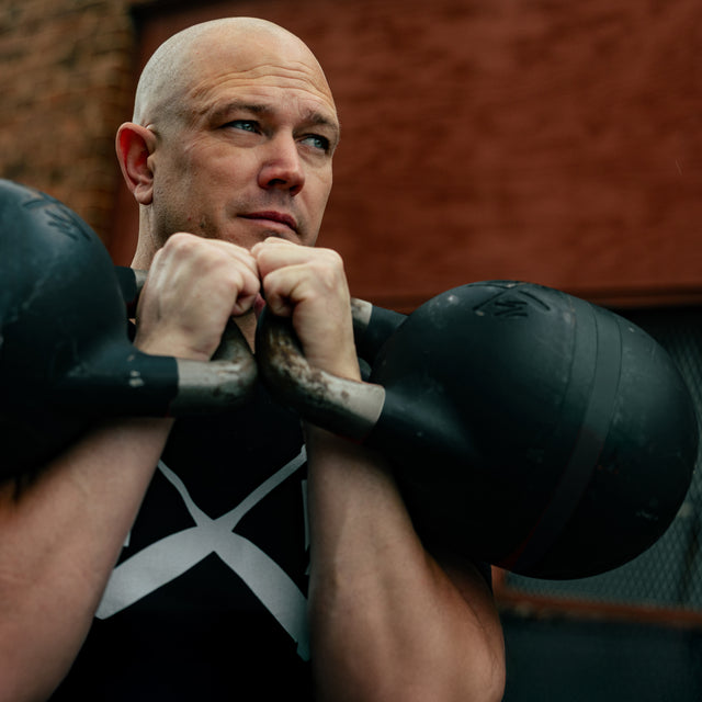 A bald man in a black shirt holds the Bells of Steel USA 48KG Extension Kit for Adjustable Kettlebell - Wildman Poison Pill at chest level, gazing thoughtfully outside with a red brick wall and metal fence behind him.