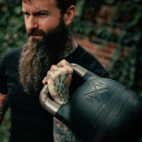 A bearded, tattooed man holds a Bells of Steel USA Wildman Poison Pill 48KG Extension Kit for Adjustable Kettlebell over his shoulder outdoors, with greenery and a brick wall blurred in the background.